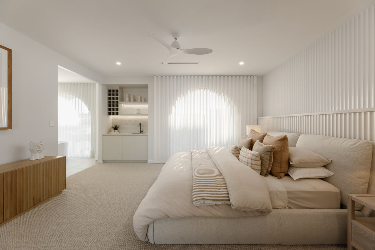 Bright contemporary bedroom featuring a curved upholstered bed, soft neutral furnishings, sheer floor-to-ceiling curtains, timber accents and a built-in kitchenette.