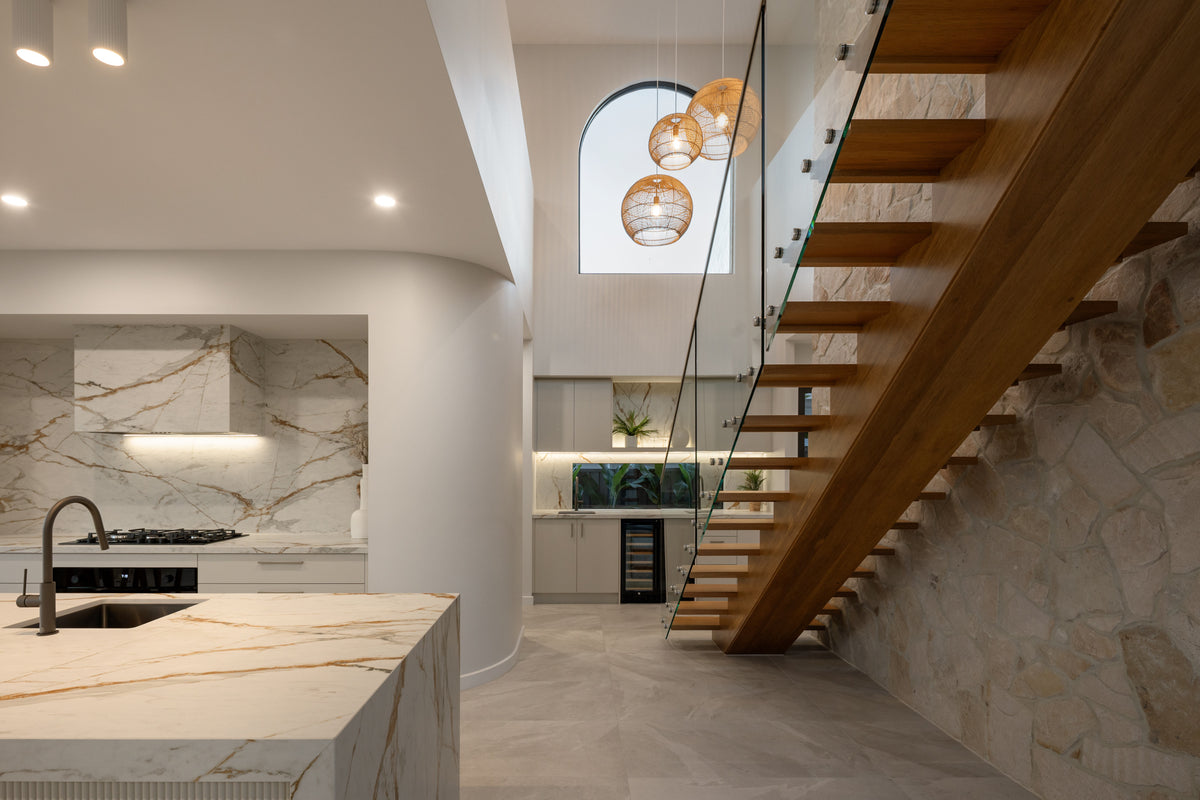 Luxury open-plan interior featuring a floating timber staircase, marble-look kitchen surfaces, natural stone wall cladding and statement pendant lighting beneath a soaring void.
