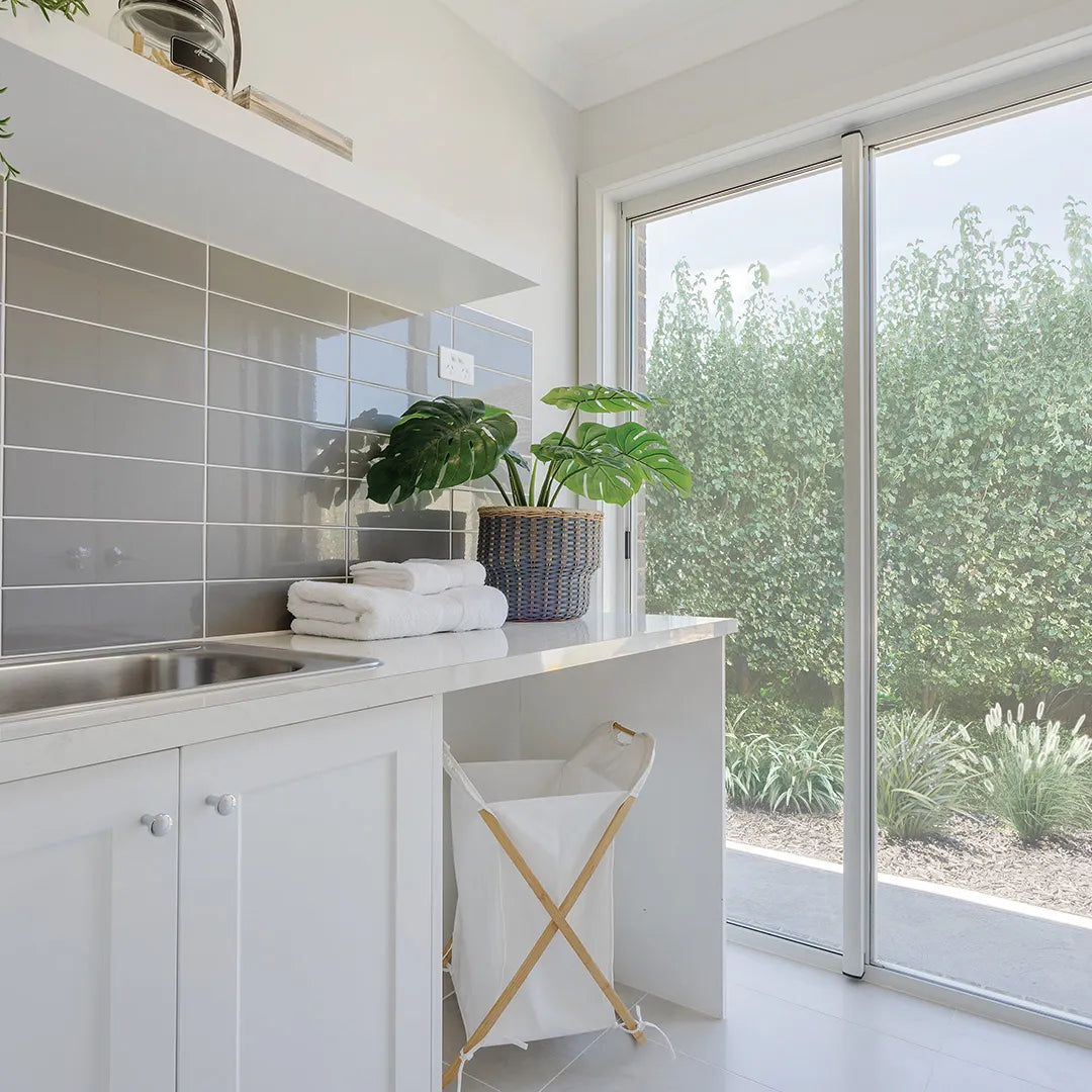 Bright laundry room featuring a glossy grey subway tile splashback, white cabinetry, a stainless steel sink, natural light and indoor greenery.