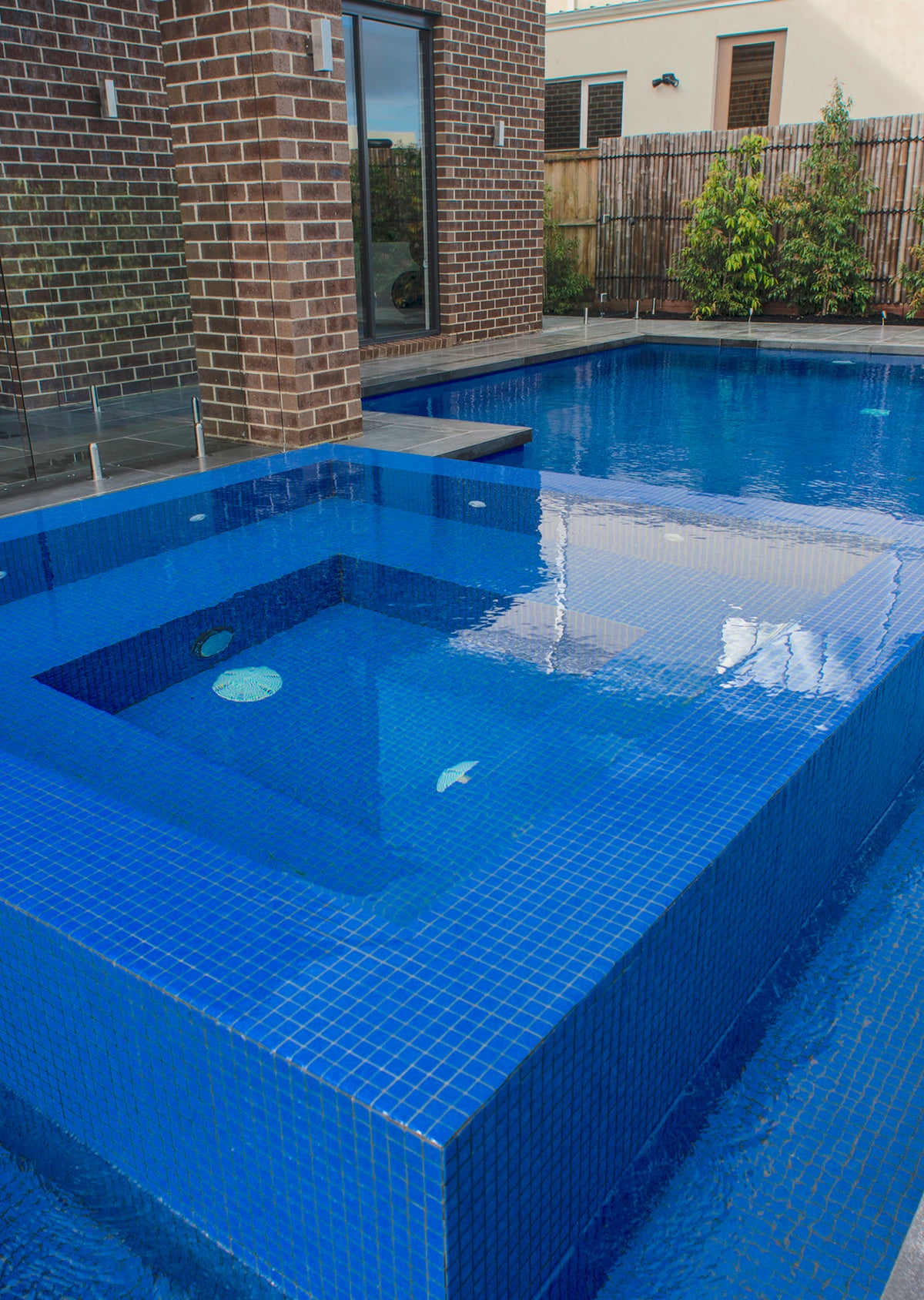 Luxury outdoor swimming pool featuring deep blue mosaic tiles, an elevated spa, glass pool fencing and contemporary landscaping beside a modern brick home.