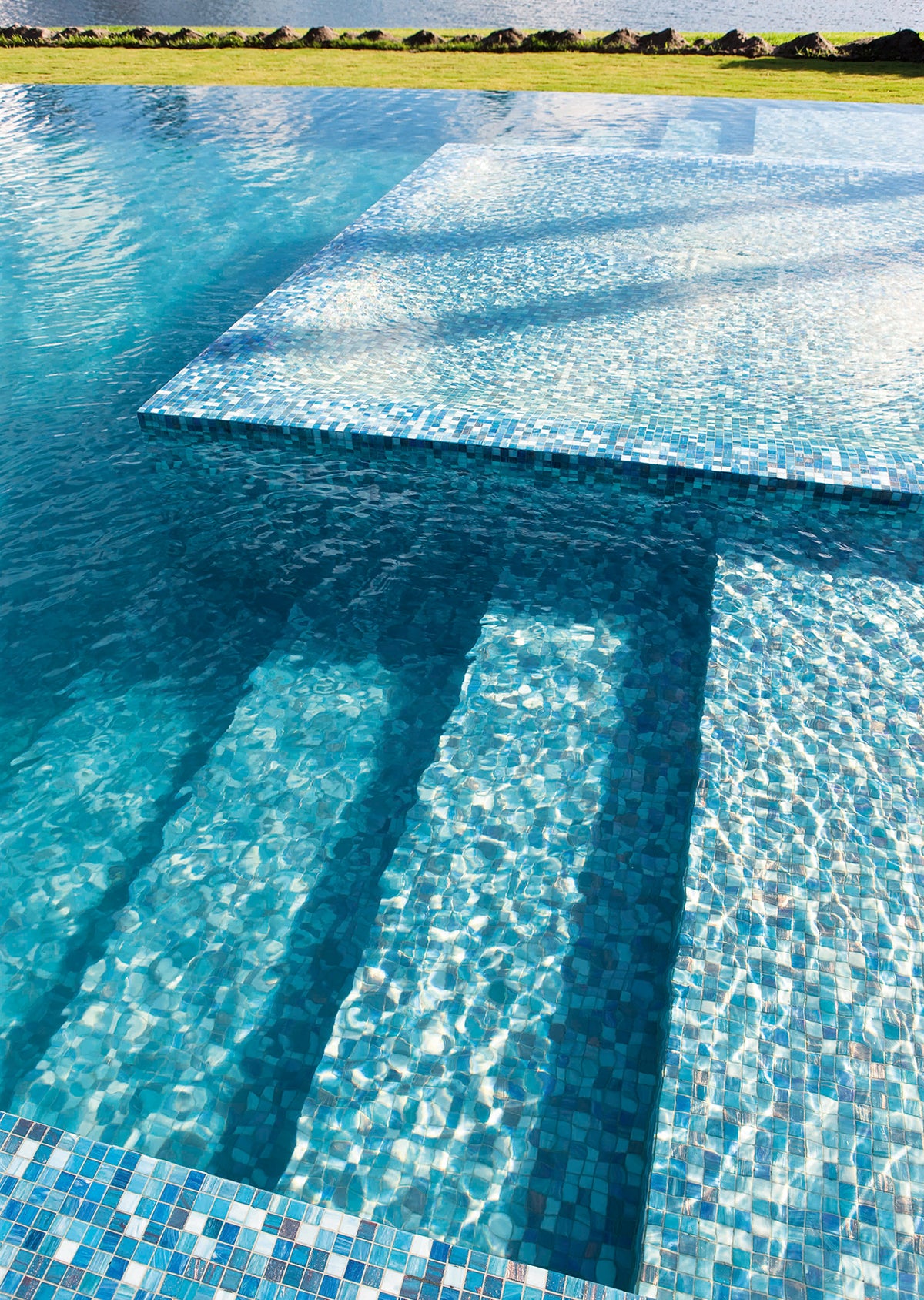 Luxury swimming pool featuring shimmering blue mosaic tiles, curved underwater steps and crystal-clear water reflecting across a resort-style outdoor setting.