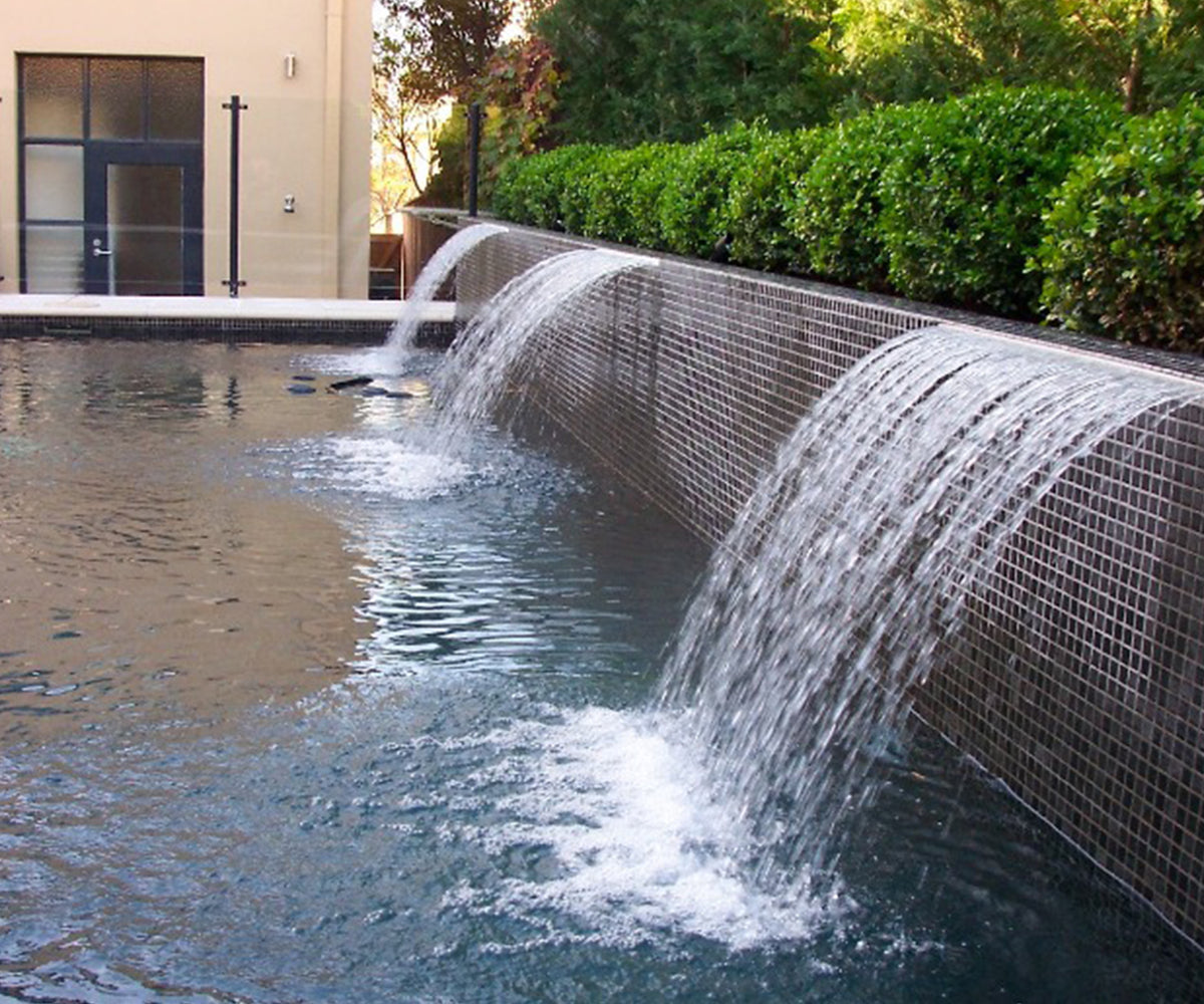 Luxury swimming pool featuring a raised wall clad in dark mosaic tiles, with three waterfall spouts cascading into the pool to create a dramatic resort-style water feature.