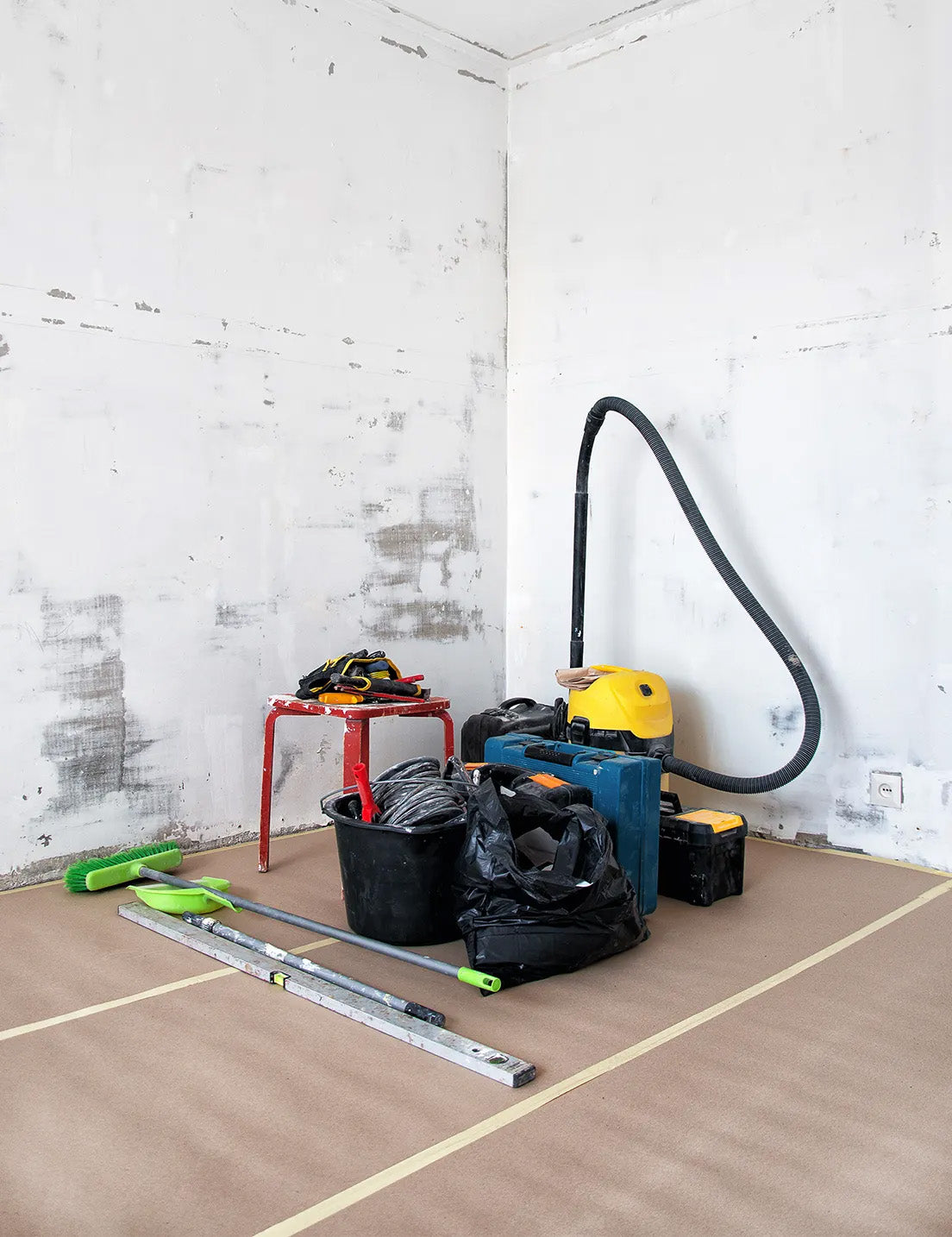 Surface preparation tools and equipment arranged in a room before tiling, including a vacuum, spirit level, broom, bucket and hand tools for floor preparation.