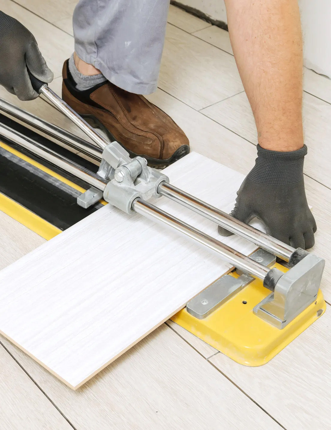 Person using a manual tile cutter to score and cut a large white ceramic tile during installation on a light-coloured tiled floor.