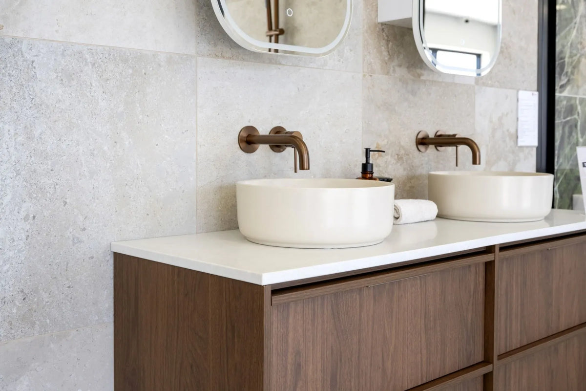 Contemporary bathroom vanity featuring twin round vessel basins, brushed bronze wall-mounted tapware, timber cabinetry and large-format stone-look wall tiles.