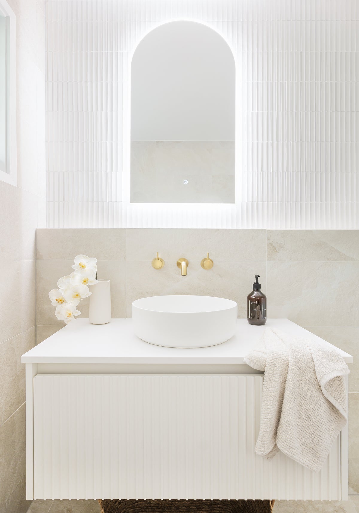 Contemporary bathroom featuring a fluted white vanity, round vessel basin, brushed brass wall-mounted tapware, an arched mirror and textured white feature wall tiles paired with soft beige stone-look tiles.