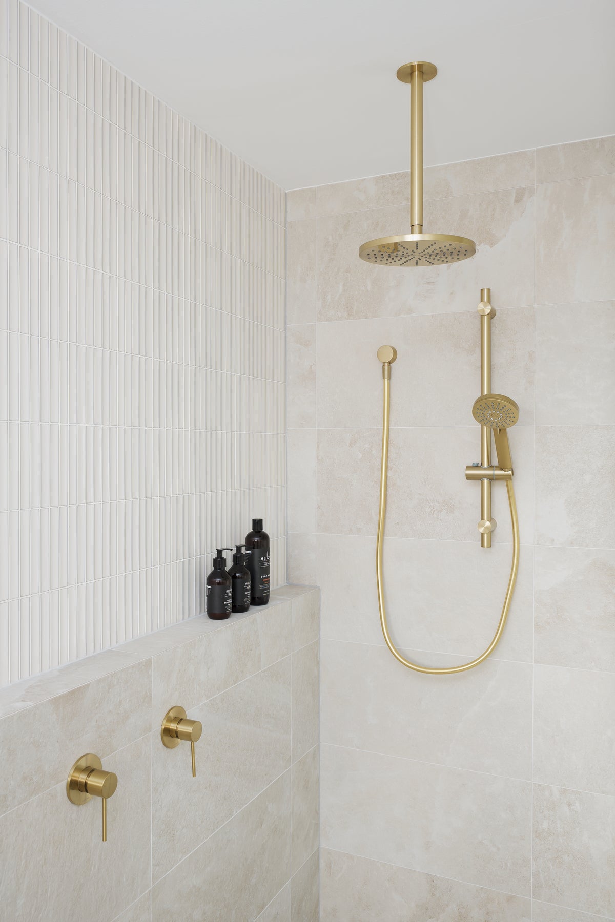 Contemporary walk-in shower featuring soft beige stone-look wall tiles, vertically textured white feature tiles and brushed brass shower fittings, including a rain shower and handheld shower.