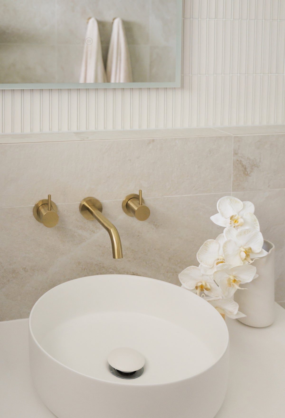 Contemporary bathroom vanity featuring a round white vessel basin, brushed brass wall-mounted tapware, textured white feature tiles and soft beige stone-look wall tiles.