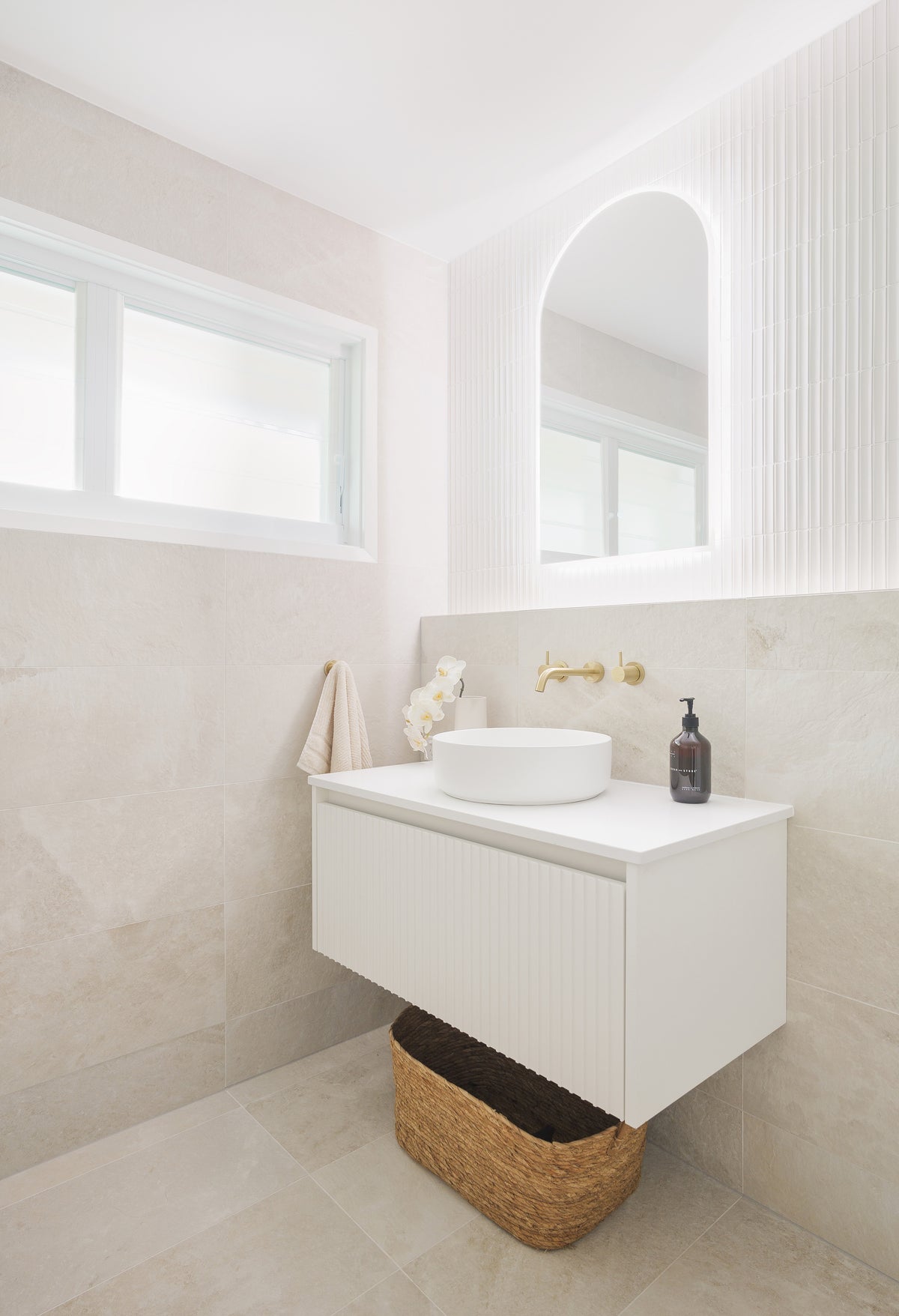 Contemporary bathroom featuring soft beige stone-look wall and floor tiles, a floating white vanity, round vessel basin, brushed brass tapware and an arched mirror framed by textured white feature tiles.