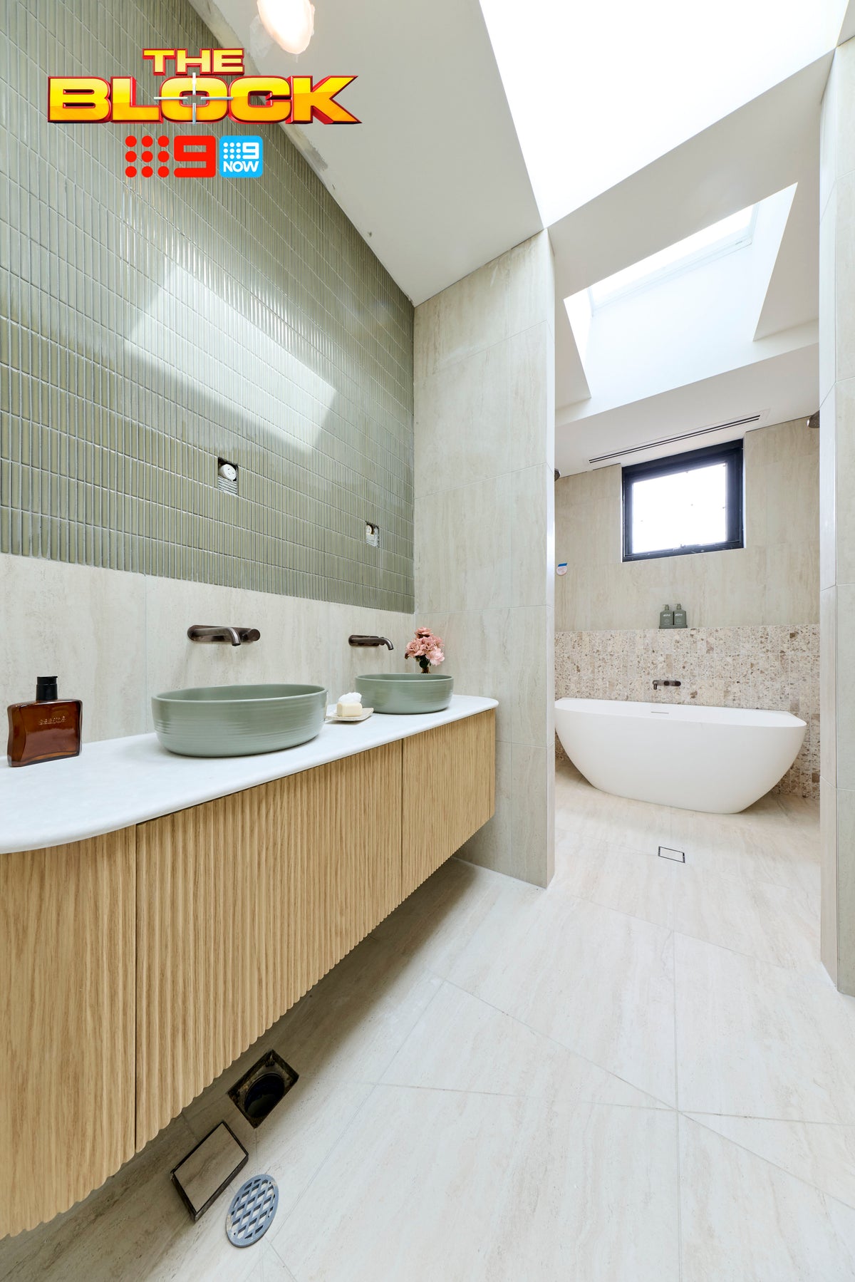 The Block House 1 main ensuite featuring soft stone-look floor and wall tiles, a floating timber vanity with sage green vessel basins, textured green feature wall tiles, freestanding bath, skylight, and pastel styling accents creating a calm spa-inspired bathroom.