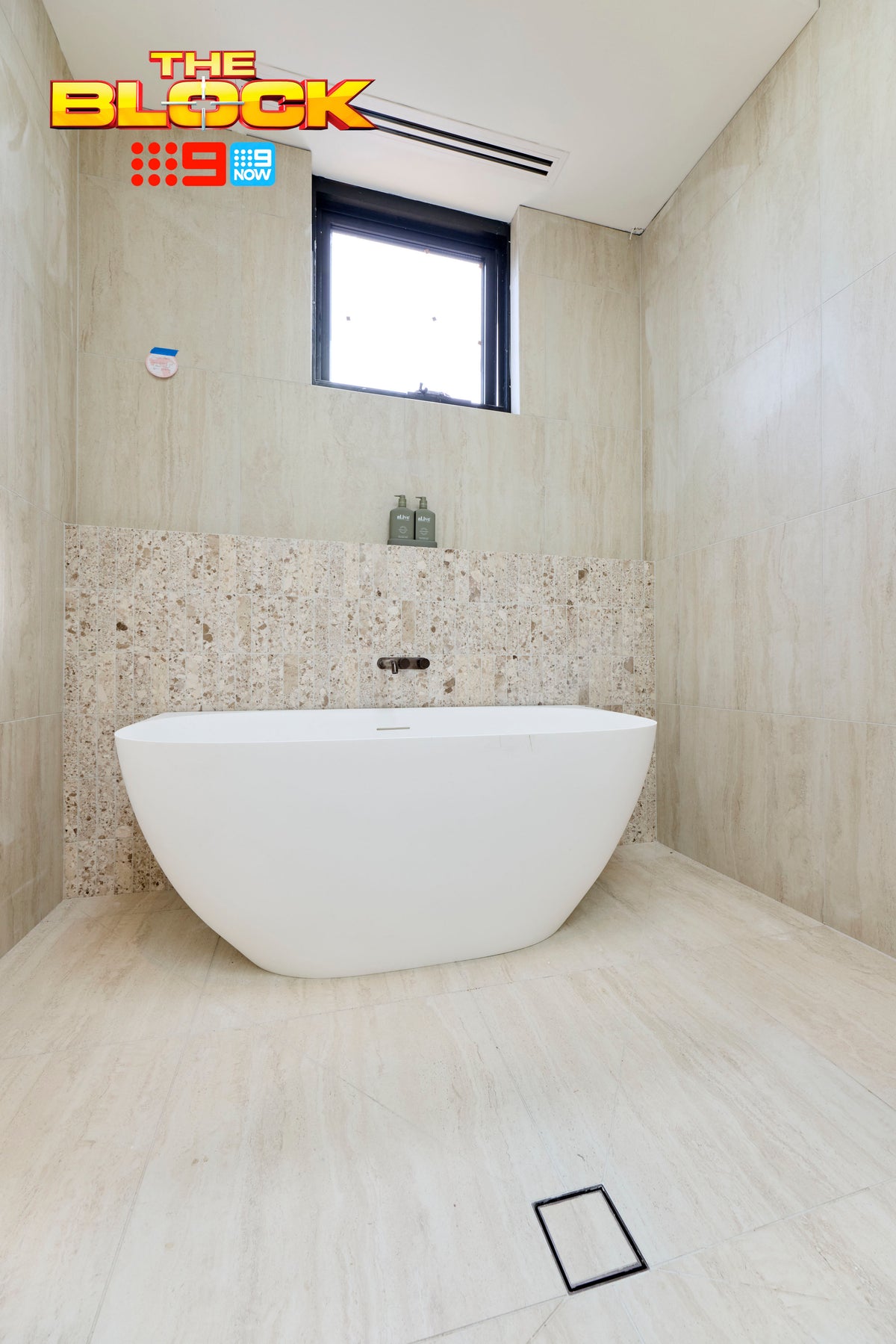 The Block House 1 main ensuite showcasing a freestanding white bath set against a textured stone-look feature wall, with large-format light beige tiles, matching floor tiles, a wall-mounted bath mixer and natural light from a high window creating a serene spa-like retreat.