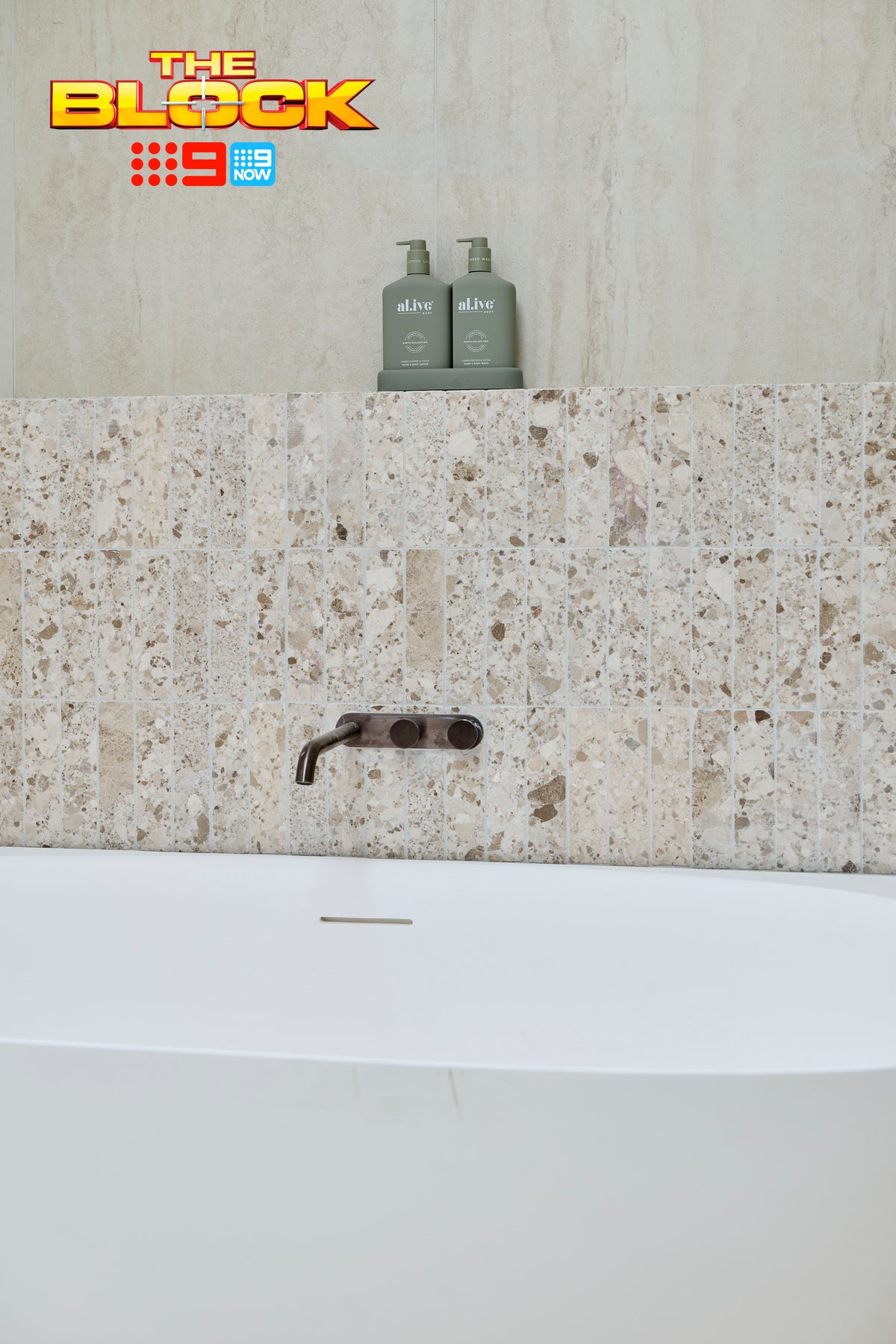 Close-up of The Block House 1 main ensuite featuring a freestanding white bath, textured terrazzo-look feature wall tiles, large-format stone-look wall tiles, and a brushed bronze wall-mounted bath mixer, creating a refined spa-inspired bathroom design.