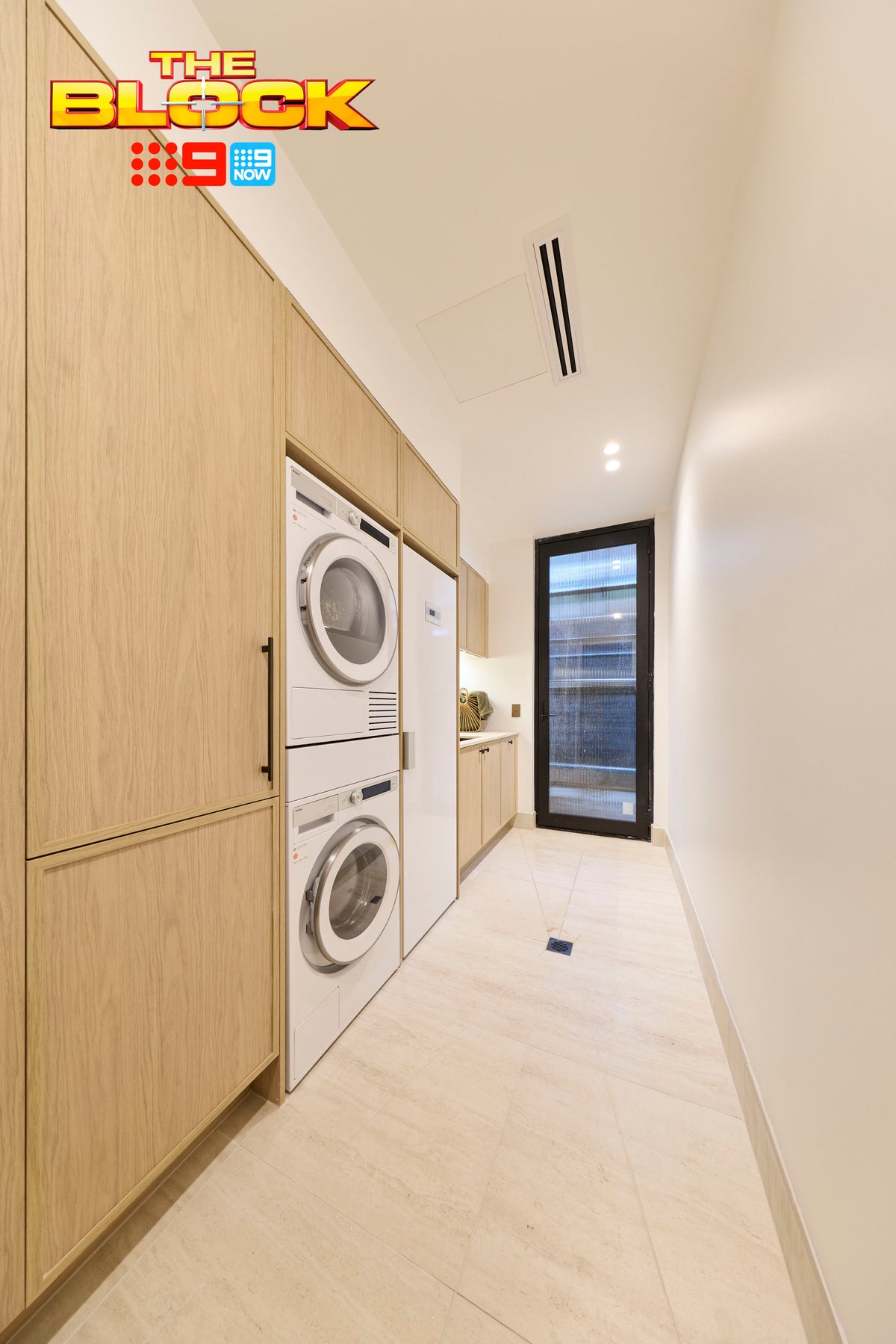 The Block House 1 laundry featuring large-format stone-look floor tiles, custom timber cabinetry, integrated stacked washer and dryer appliances, a built-in utility area, and a sleek contemporary design with warm natural finishes.