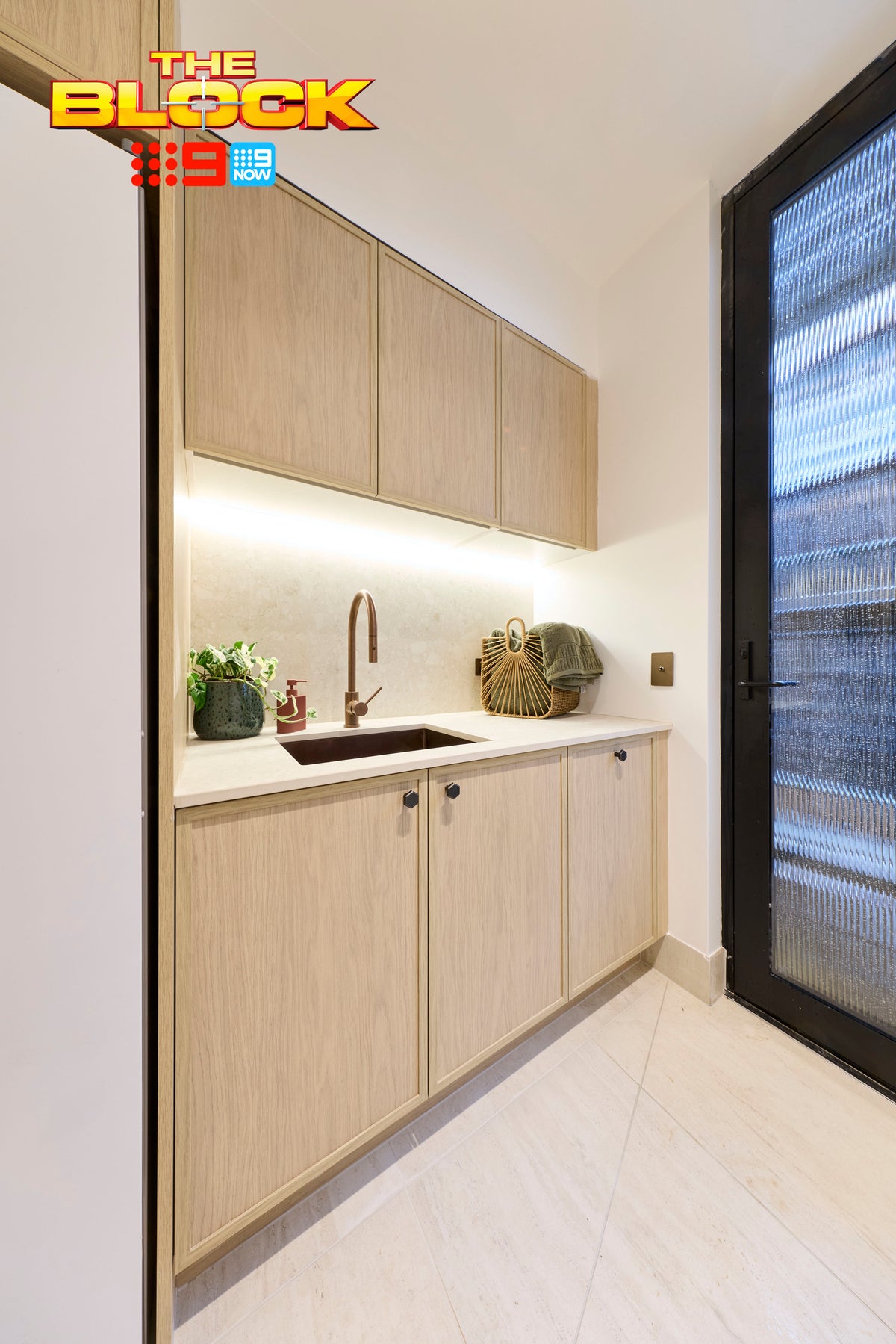 The Block House 1 laundry featuring custom timber cabinetry, a stone-look benchtop and splashback, integrated sink with brushed bronze tapware, large-format stone-look floor tiles, and a frosted glass external door for natural light and privacy.