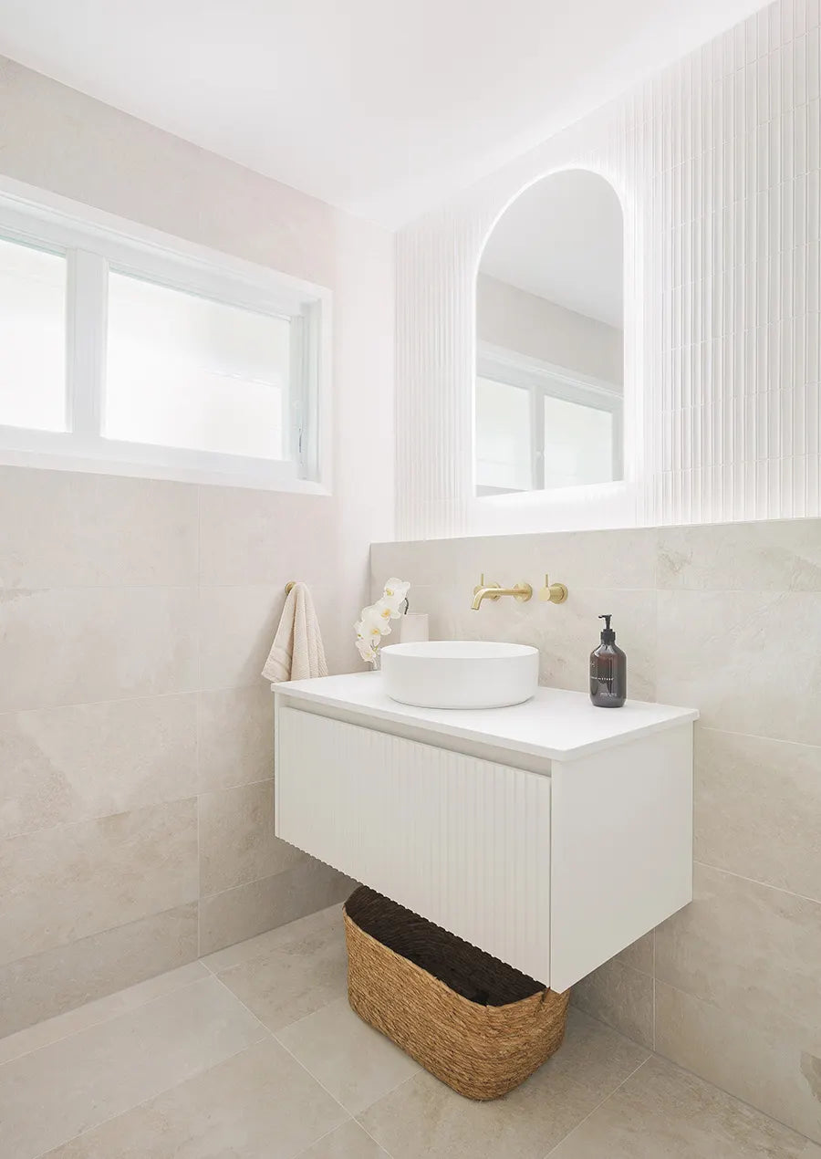 Contemporary bathroom featuring soft beige stone-look wall and floor tiles, a floating white vanity, round vessel basin, brushed brass tapware and an arched mirror framed by textured white feature tiles.