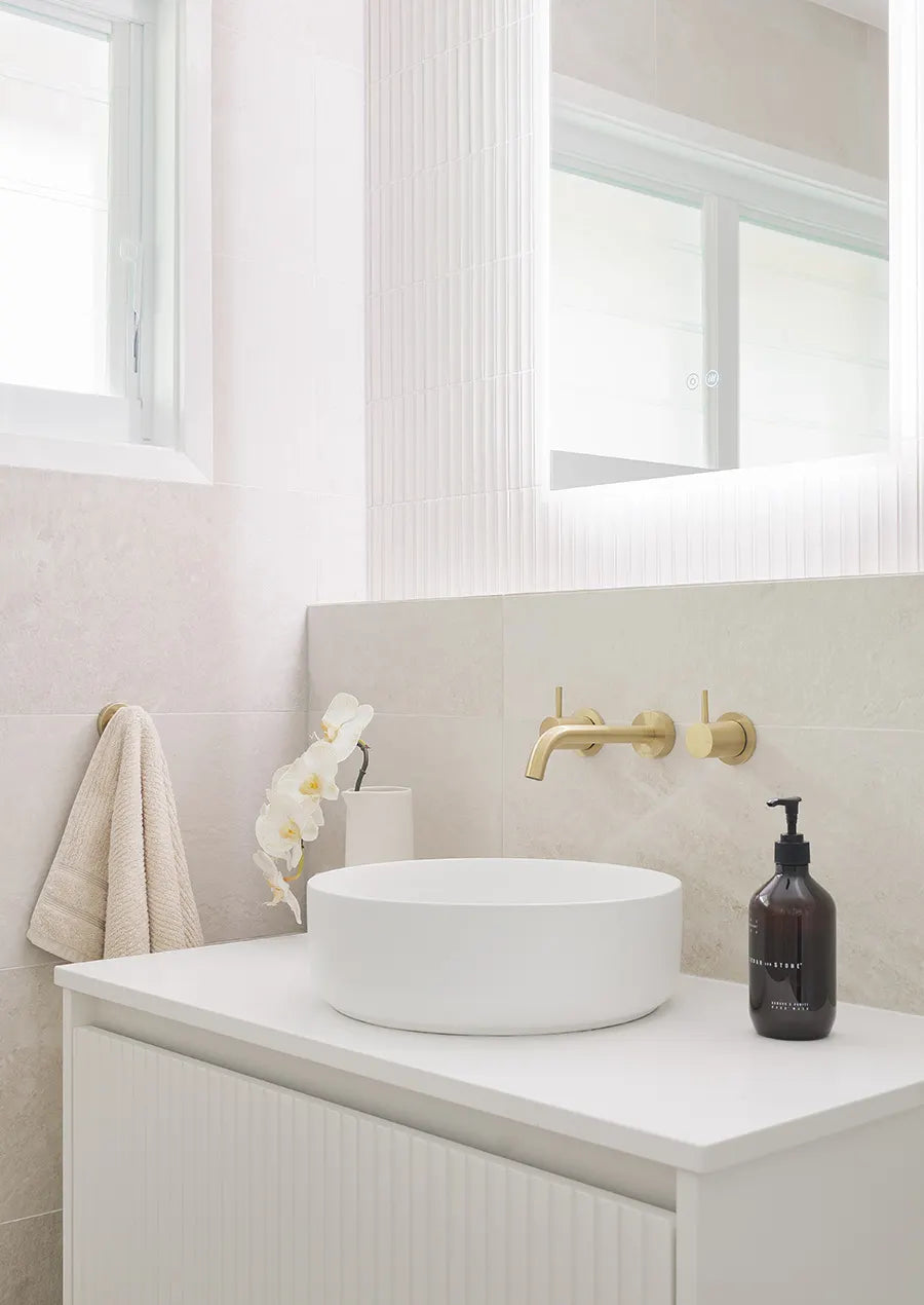 Contemporary bathroom vanity featuring a round white vessel basin, brushed brass wall-mounted tapware, textured white feature tiles and soft beige stone-look wall tiles.