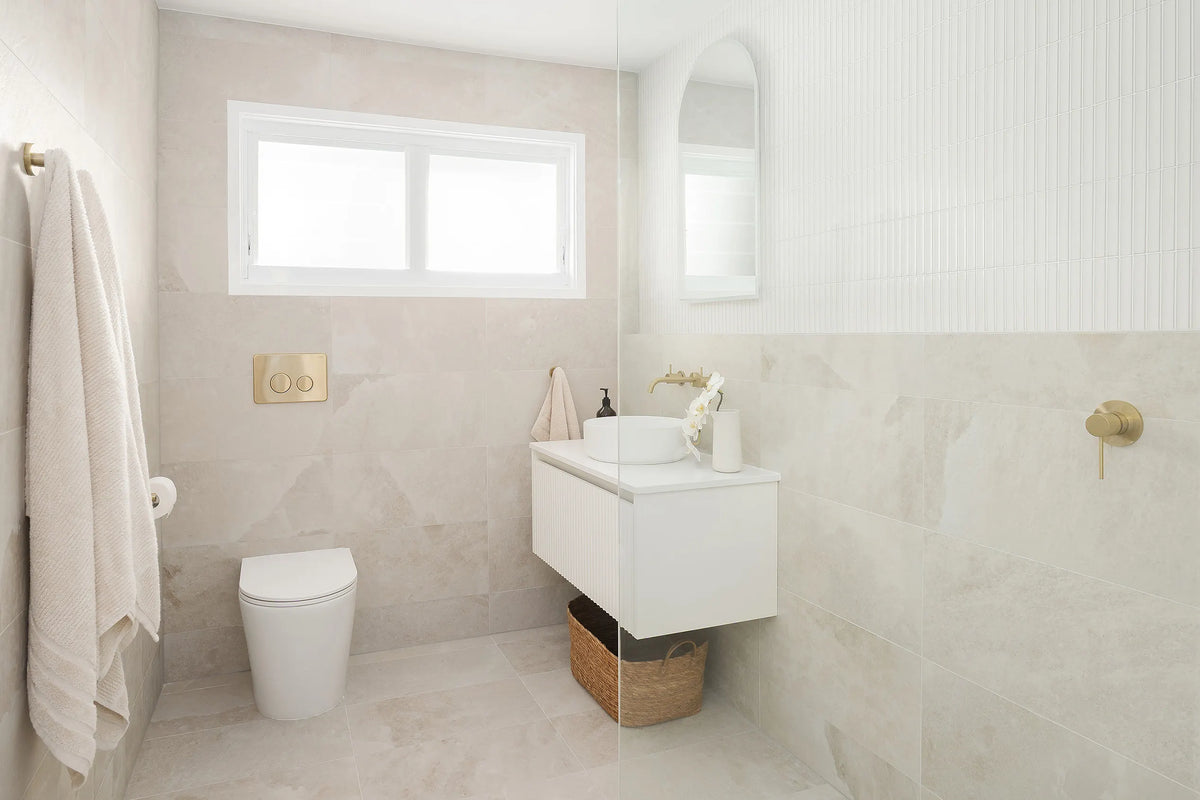Contemporary bathroom featuring soft beige stone-look wall and floor tiles, a floating white vanity, round vessel basin, brushed brass tapware and an arched mirror framed by textured white feature tiles.