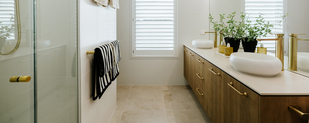Modern bathroom with a timber double vanity, brushed brass fixtures, frameless shower and neutral stone-look floor tiles.